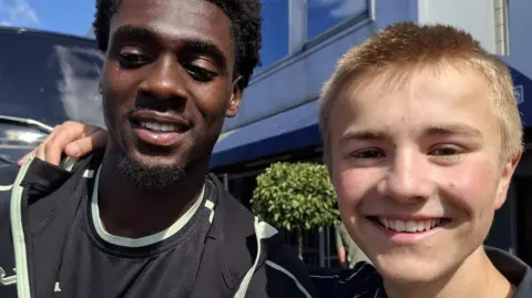 Norwich striker Mo Touré (left) is wearing a black T-shirt. He is standing next to Ranulph Bacon, who is wearing a navy polo shirt. The pair are smiling at the camera.