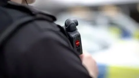 A close up of a police officer's shoulder. They are wearing a bullet proof vest with a radio mounted on it. A police car is blurred in the background.
