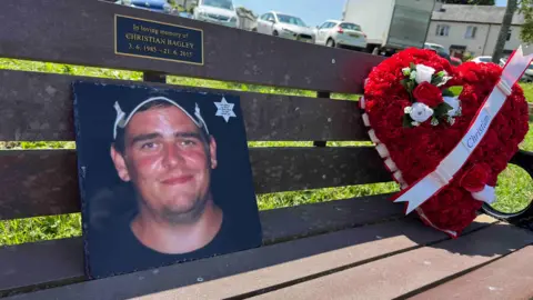 A wooden bench with a photograph of a young man wearing a baseball cap resting against the backrest. A bouquet of red flowers in the shape of a heart is also on the bench. A ribbon across it reads "Christian". A plaque on the bench reads: "In loving memory of Christian Bagley - 3.6.1985-21.6.2015".