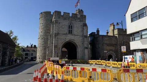 A stone tower with a large archway underneath it, flying a union jack flag from its turrets. Road access to the archway is blocked by orange traffic cones and yellow plastic barriers.