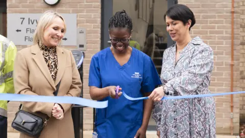 Three women stand alongside each other to cut a ribbon unveiling the new housing. In the centre is a nurse called Dorcas, who is smiling and cutting the light blue ribbon with a pair of scissors. She is wearing blue nurses scrubs. The women either side of her are smiling and one is holding the ribbon.