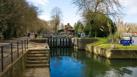 A lock with an area of grass to the right and a walkway to the left. There are trees in the background. 