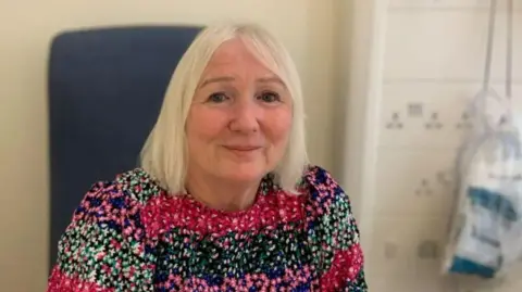 Lesley Harrison smiles nervously at the camera as she sits in a hospital chair 