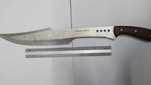 Lincolnshire Police A large kitchen knife with a brown handle and a curved, metal blade, which shows signs of wear and discolouration. It is shown next to a 30cm ruler and is almost twice as long