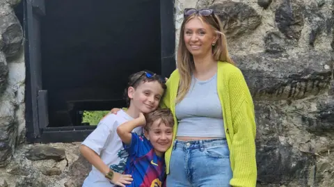 Zoe Stokes-Jones Photograph of Zoe Stokes-Jones (right) pictured with both of her sons. Jacob is on the left, with her youngest son in the middle. She wears a bright green cardigan, a grey t-shirt and blue jeans. She has blonde long hair and sunglasses on her head. They stand outside a stone wall building. 