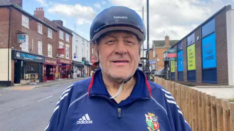 A man stood down a road, wearing a blue sports jacket and a cycle hat.