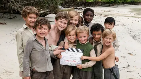 Alfie and his fellow Littleuns in character on the beach, holding a clapper board