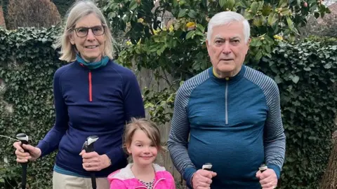 Supplied Two grandparents, a man and a woman, wearing walking gear and holding walking poles. Their granddaughter, wearing a pink coat, is stood in between them.