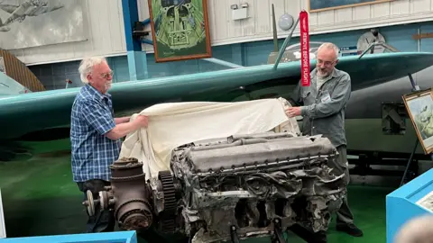 George Carden/BBC Two museum staff remove a sheet off the spitfire engine inside one of the hangars. Behind them is a sky blue spitfire wing