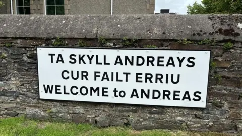 EMMA DRAPER A large white sign that says Welcome to Andreas in English and Manx in black lettering. it is attacked to a stone wall and there is a building in the background and green grass in the fore.