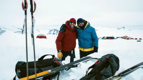 National Geographic/Freddie Claire Will Smith and Richard Parks are hugging shoulder to shoulder wearing thick winter coats in front of snow exploring gear. They are stood in a snowy environment in Antarctica. 