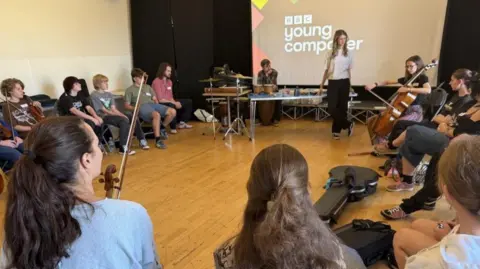 A dozen teenagers - a mix of boys and girls - seated in a circle. Some are holding musical instruments, including violins and a cello. In the centre a young woman is standing next to a table and a set of drums, with a backdrop projection stating BBC Young Composer