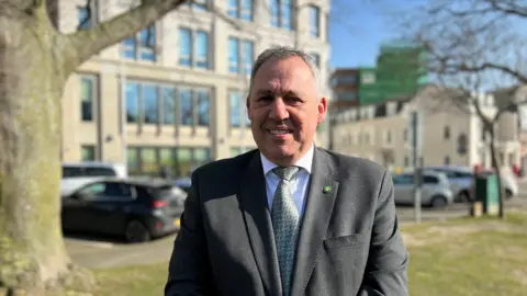 Andy Jehan seen wearing a grey suit with a white shirt and green patterned tie. He has short grey hair and is stood outside near a street with cars and a large tree to his right. 