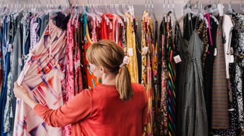 A stock image of a woman looking through a rail of brightly-coloured clothing in a charity shop. 