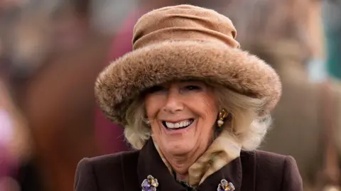 PA Queen Camilla smiles at the camera as she stands in the parade ring at Cheltenham Festival. She has short blonde hair and wears a light brown fur hat, a burgundy tailored jacket with floral broaches on the lapel. She wears gold earrings.