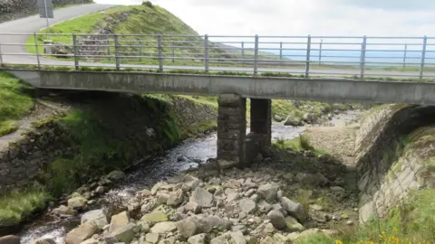 Buckbarrow Bridge, which crosses a small stream. It joins two roads and is in a rural location. There are rocks on under the bridge, which is supported by two pillars made from stones.