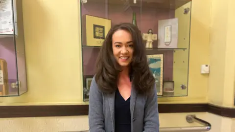 Woman with long brown hair and grey blazer and black top stood in front of yellow wall