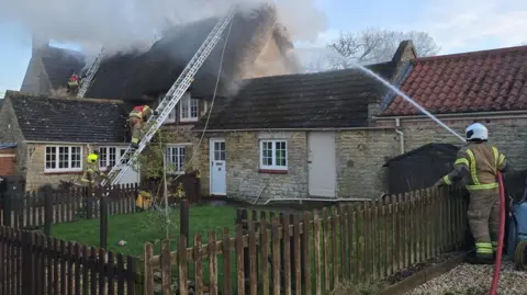 NFRS Fire crews on ladders putting out fire in rooves. One crew member is holding a jet hose which is spraying water at a thatched roof. There is a lot of smoke. 