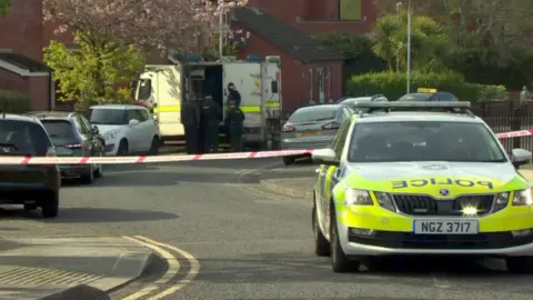 A police car is parked in front of a white and red cordon in a residential street. Multiple cars are parked outside houses on either side of the street. In the distance five police officers are standing beside a white police van. 