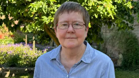 A woman with dark hair and glasses is looking at the camera. She is wearing a blue top. She is standing in a garden with a stone wall, surrounded by trees and flowering plants in the background.