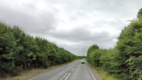 Street view image of the A602. It is a long straight road with bushes on either side. There is a car heading towards the camera. There are two lanes in the road - one for each direction. 
