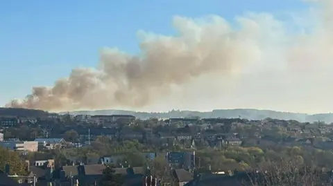 City rooftops with smoke rising up from a hill in the background against a blue sky
