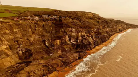 Jamie Woodford/ Wight Eagle Drone Services A brown crumbling cliff with the sea visible below and the amusement park on the top.