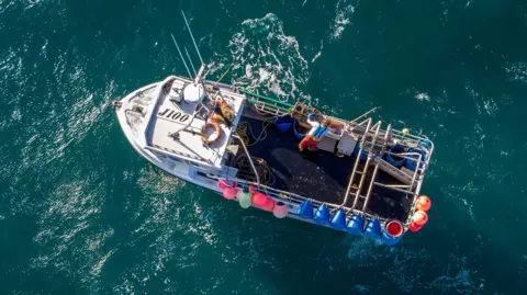 An overhead shot of a man working on a fishing boat in the North Sea. He is wearing a bright-coloured top and red trousers and is standing on the right of the boat.