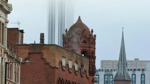 A gothic red brick building with a high decorated dome shaped roof has been pictured with light amounts of smoke coming from the top of the building. 