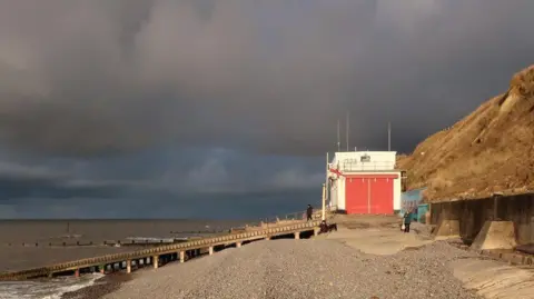 Geograph/Hugh Venables Lifeboat station in white and red is perched underneath a grassy cliff on the sea edge. A wooden ramp leads from the lifeboat house to the sea. The scene is moody with a dark sky and sunshine. Dogs and owners can be seen.