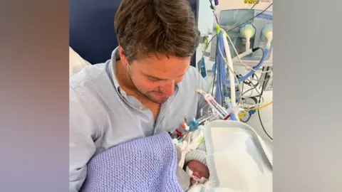Nick and Libby Powell Nick Powell is pictured holding his son. The picture is taken from above and he is looking at his baby who is covered in a blue blanket. The baby has a woollen hat on and there are tubes on his face and around him. They are in a hospital setting.