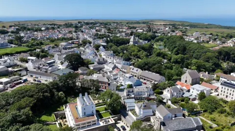 BBC An aerial view of St Anne in Alderney. It shows rows of houses and shops form an elevated view with the sea in the background and plenty of trees and fields. 