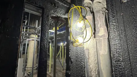 Shaun Whitmore/BBC The inside of the kitchen where a fire started at St John the Baptist Church in Harleston. The walls have been charred and there is nothing left of the doors. Through the gap where the door previously was, scaffolding has been put up to give access to higher parts of the church. A light has been strung up with a yellow wire.
