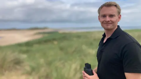 A man with short blonde hair, wearing a black T-shirt, holds a pair of black binoculars on a grassy beach dune.