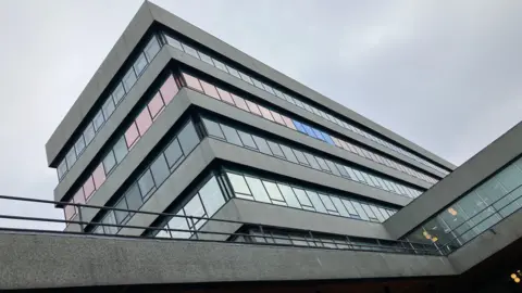 A six-storey rectangular office block with bands of glass windows, separated by grey concrete panelling.