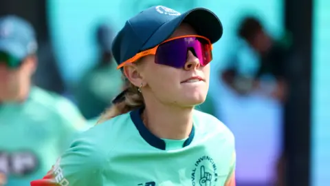 Getty Images Sophia Smale wearing a fluorescent green top, orange sunglasses and a navy cap while playing cricket.