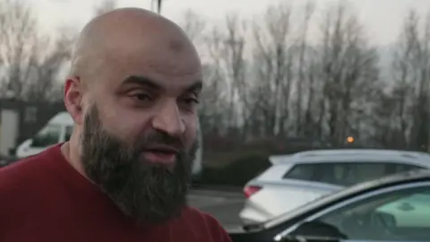 A man with a dark beard and shaven head stands in a car park. He wears a red sweater.