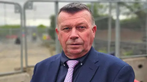 Peter Osborne in a suit and tie, looking at the camera. Construction fencing and road works can be seen in the background.