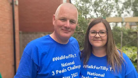 BBC / Shirley Henry Stuart and Pam Thistleton wearing blue Walk For Tilly T-shirts. Pam has brown shoulder-length hair and wears glasses. Stuart has shaved grey hair