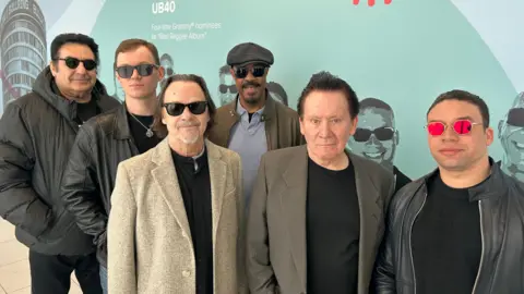 BBC Six men lined up against a blue wall. Five out of six wear sun glasses while one is without wearing a grey blazer on top of a black shirt.