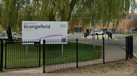 The exterior of the school from behind it's black gates. A purple and white sign on the field reads: "Welcome to Grangefield Academy". Down the driveway to the building is a car park. The orange brick building is long and three-storeys tall.