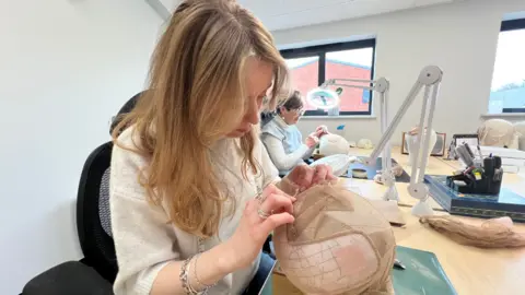 A woman making a wig on a modelling block covered in a fine mesh. The wig stylist has long blond hair and wears a white jumper and jewellery. She is in a room with other stylists.