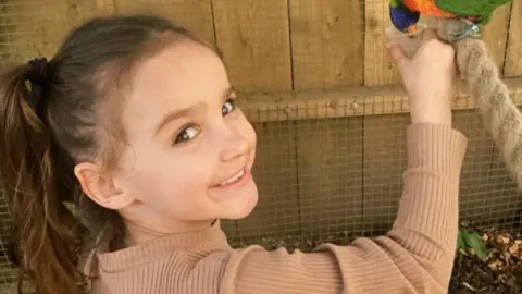 Family handout Aria Thorpe. She is a young child with long, dark hair tied up in a ponytail. She is pictured feeding a colourful bird and is looking over her shoulder to smile at the camera.