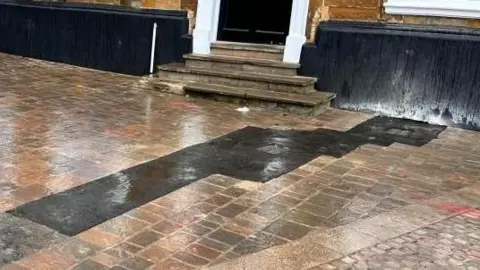 A long, black asphalt strip strip runs over some new-looking brick paving. The paving is in front of an old, stone building with a black door.