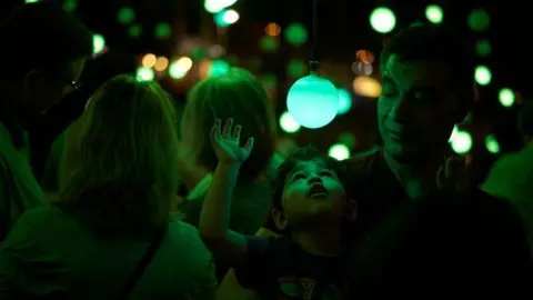 Gloucester City Council A small child, held by an adult, looks up in amazement at a blue glowing bulb as part of an art installation. There are other people out of focus in the background, also looking up at dozens of blue, green and orange glowing bulbs suspended from the ceiling.