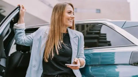 A woman is standing by a green car with the door open. She has a mobile phone in her hand. She has long blond hair and is wearing a light blue jacket and a black top and is looking happy.