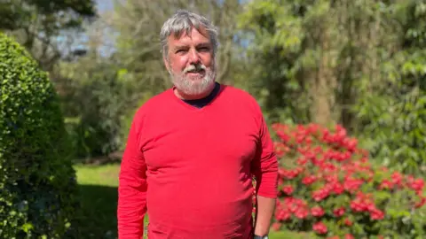 Deputy Peter Roffe has a grey beard and short grey hair and is wearing a red T-shirt standing in a garden