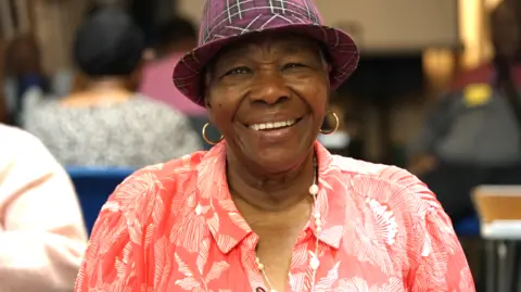 A black women wearing a red and white patterned blouse and purple plaid trilby hat smiles into the camera