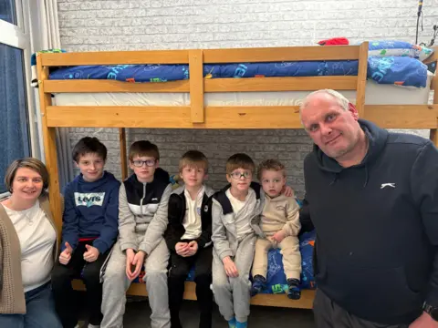 Mother Sam stands to the far left wearing a white T-shirt, cardigan and jeans. She has a short bob hairstyle. Next to her, sitting on the bottom bunk bed are her five sons all lined up together wearing tracksuits. Their father Jason is standing on the right wearing a navy hooded top. He has short grey hair.