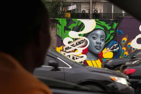 Sodiq Adelakun / REUTERS A man in a car looks past other cars at a brightly coloured mural of a woman and a peacock.
